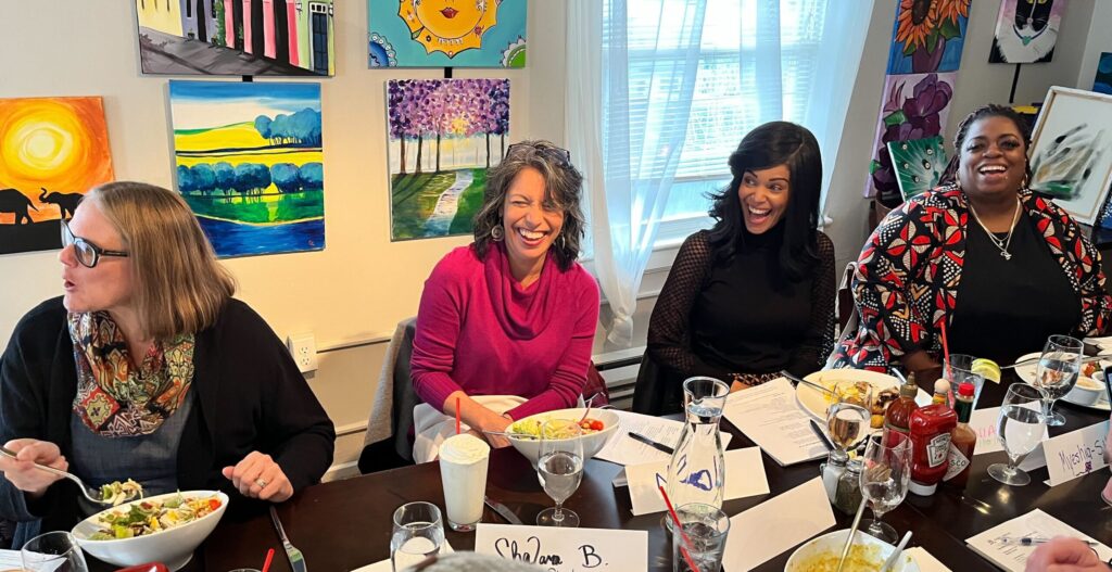 Four of Magic Cabinet's Tacoma 2 Cohort members are all smiles while sitting at a table with art on the walls in the background.