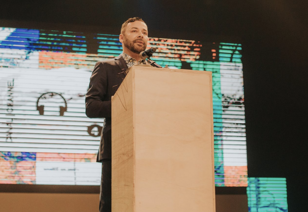 A man wearing a black suit and patterned shirt stands at a podium and prepares to give a speech.