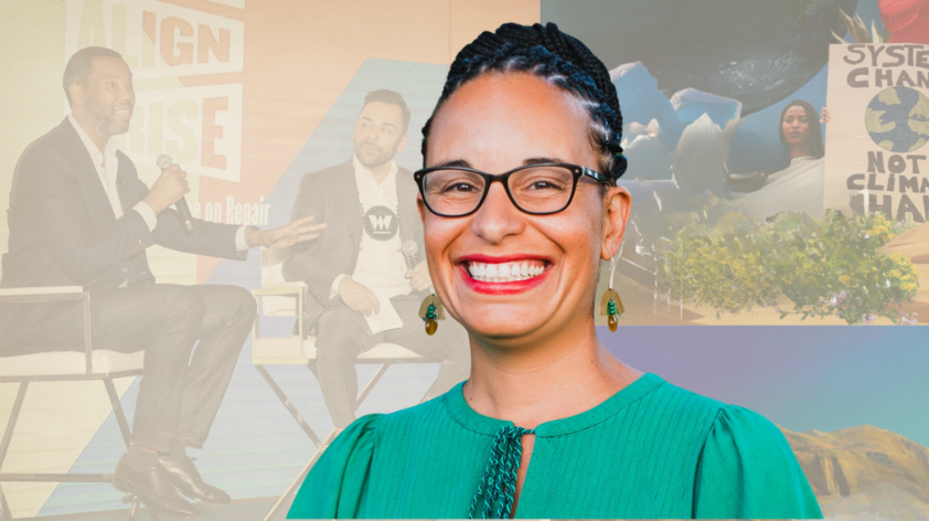 A woman wearing a green top and glasses with braids in her hair smiles against a transparent backdrop of two speakers at a conference.