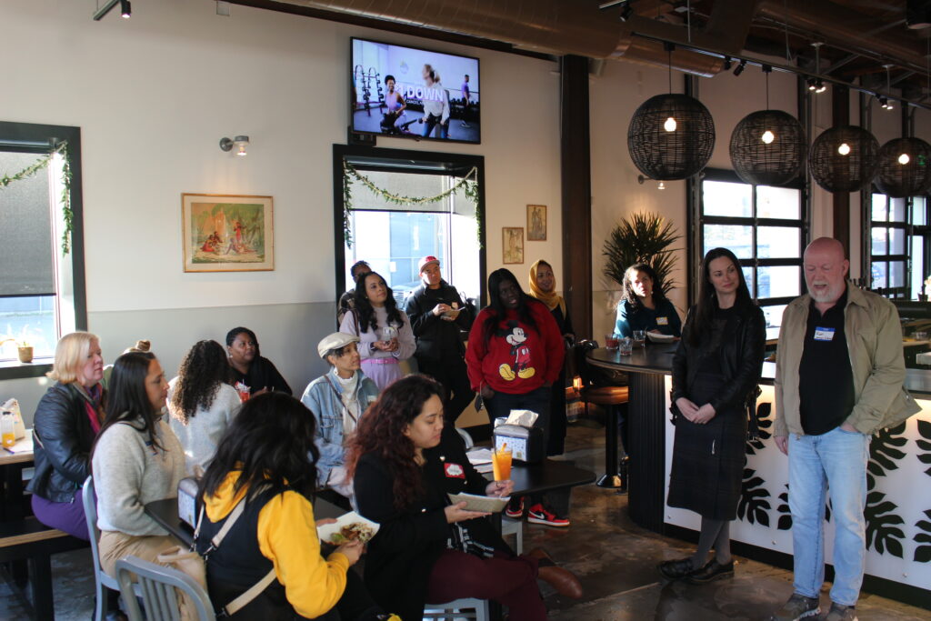 A group of people are gathered in a restaurant space. A man and woman stand at the front of the room making speeches while everyone looks on.