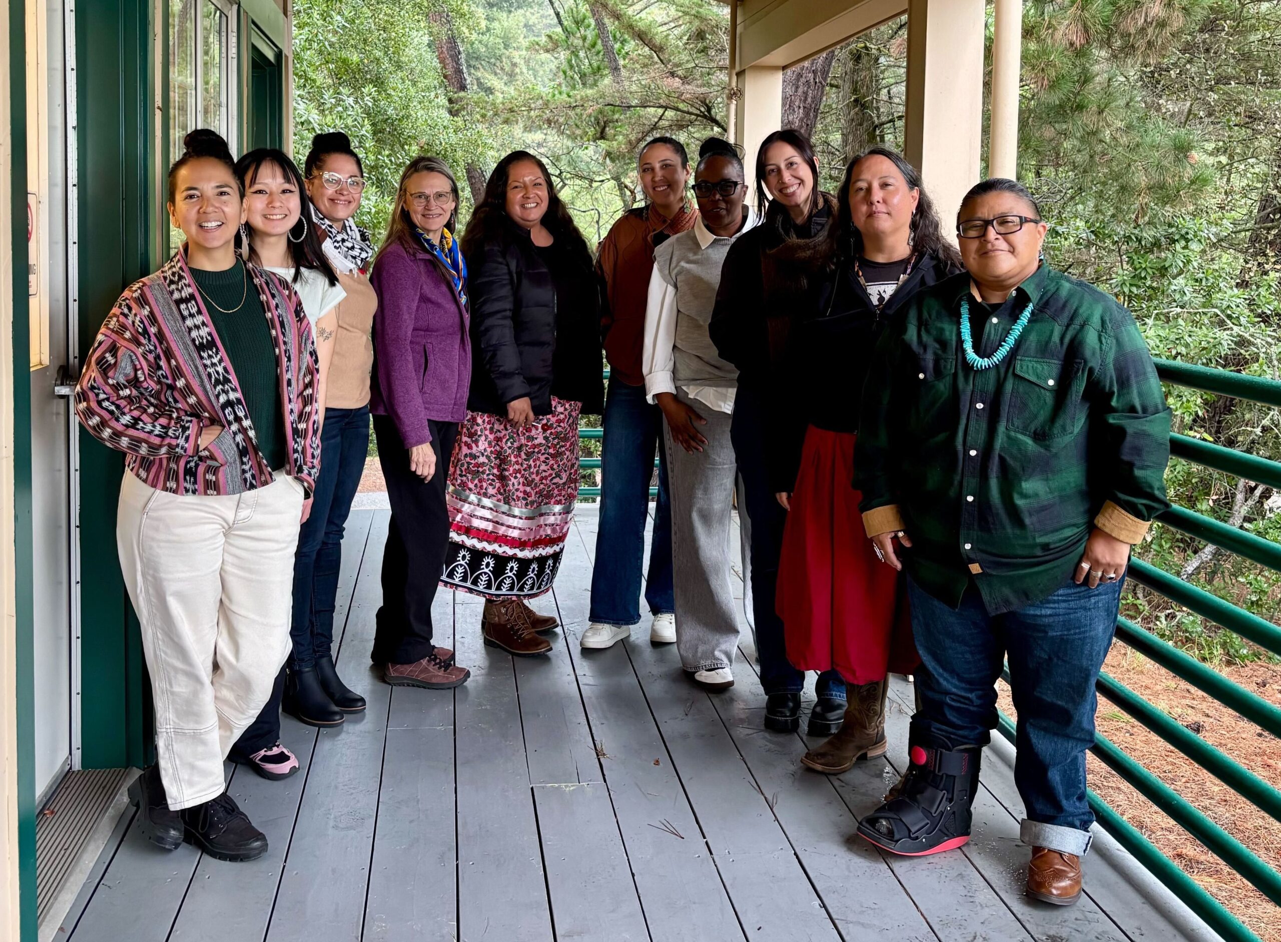 A group of people stand in a semi-circle on a porch with a wooded area in the background.